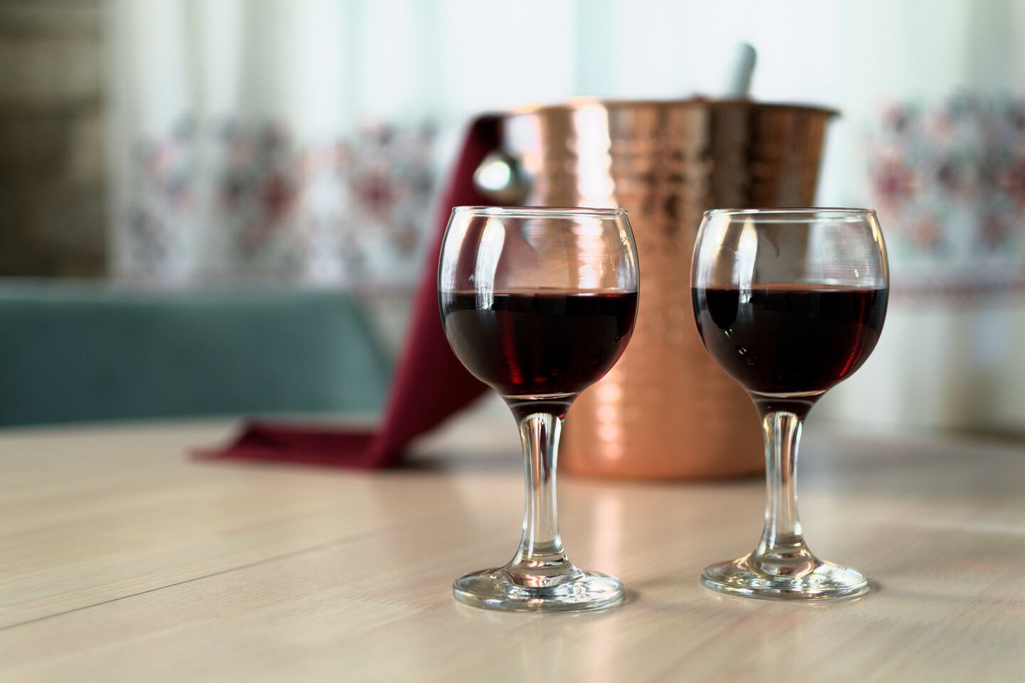 Dois copos de vinho tinto sobre uma mesa de madeira, com uma panela de cobre ao fundo.