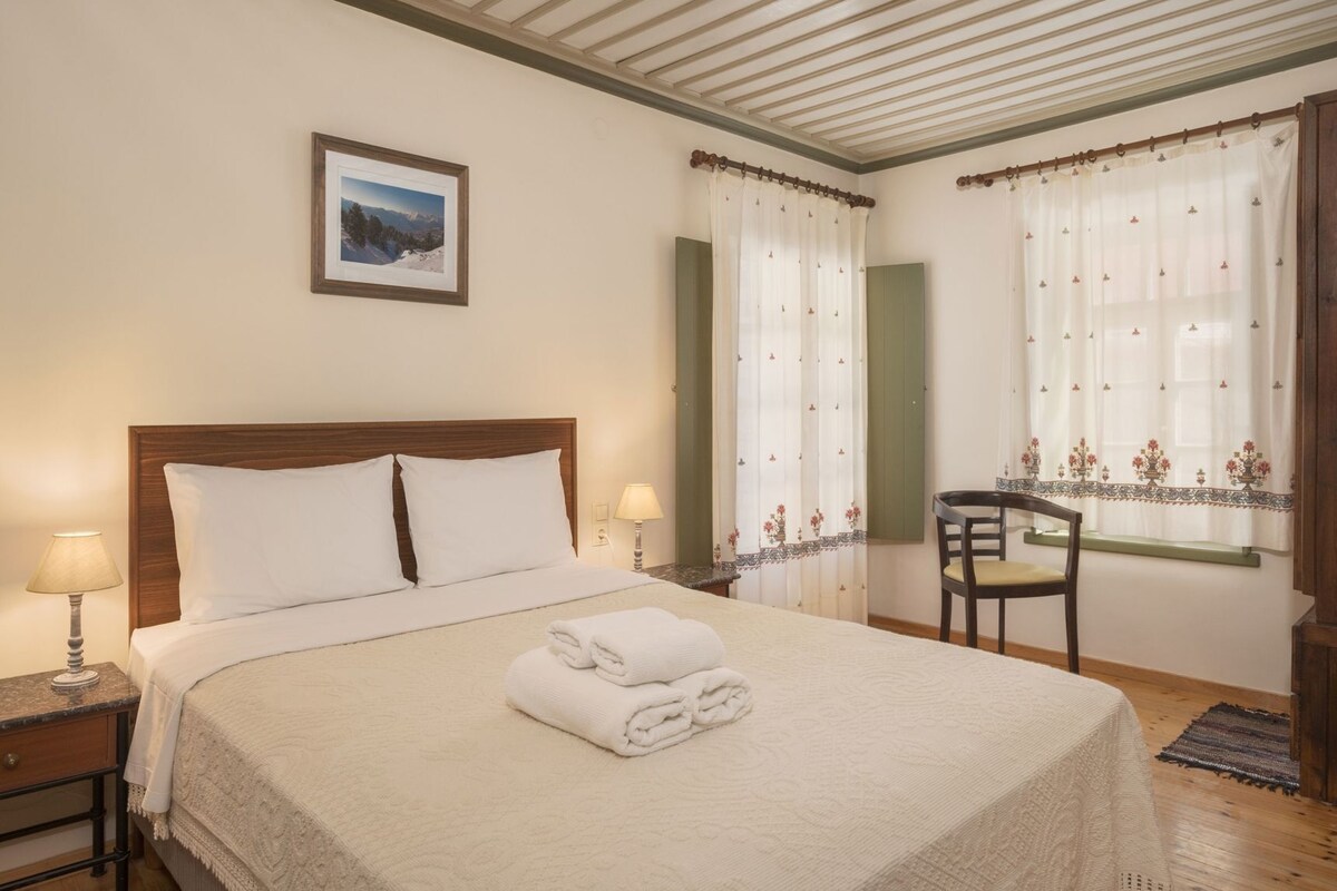 A hotel room with a bed, two bedside lamps, a chair, a window with curtains, and a framed picture on the wall.