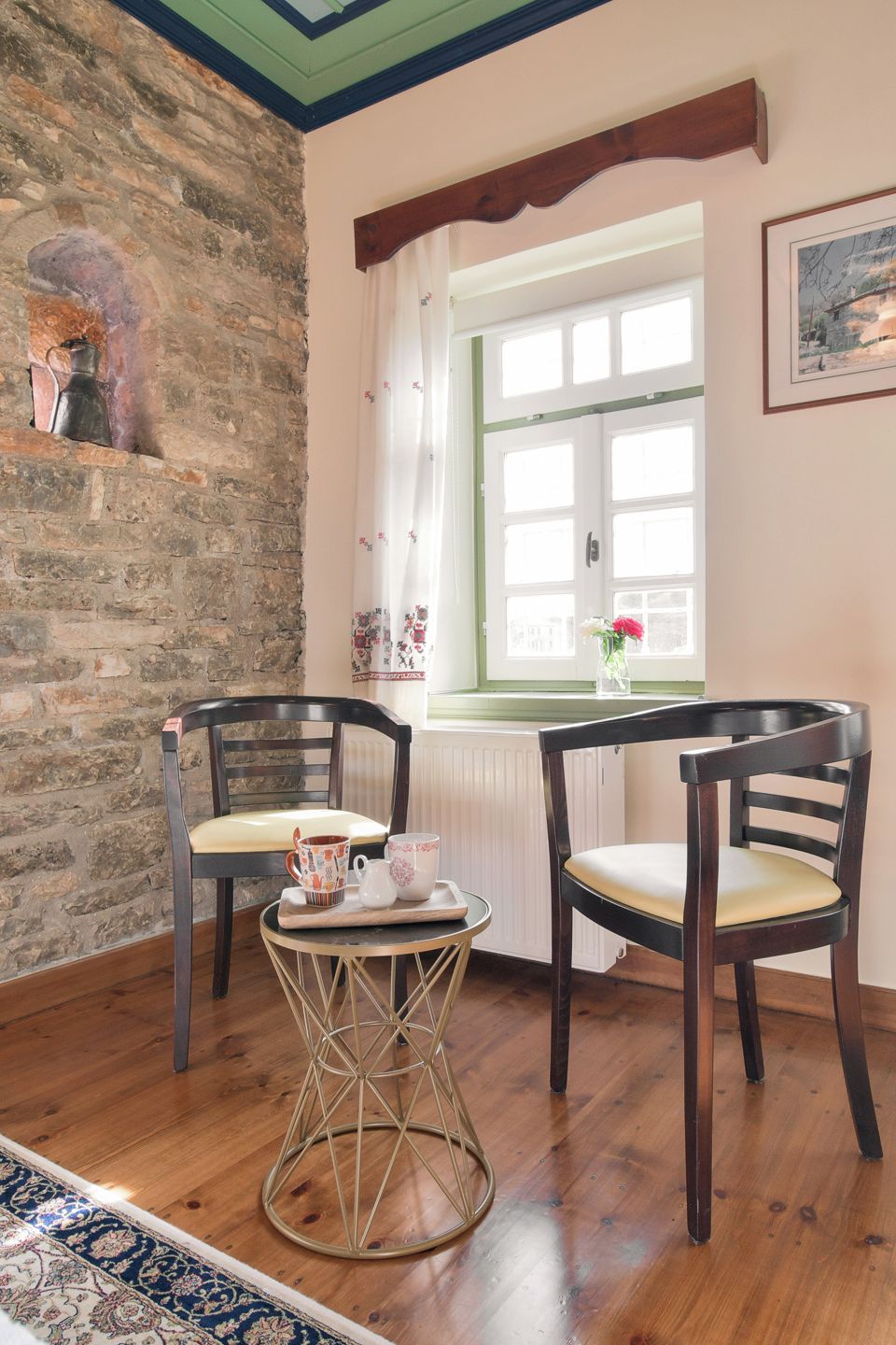 Una habitación acogedora con pared de ladrillo, dos sillas de madera, una mesita redonda con una bandeja y una ventana con cortinas.