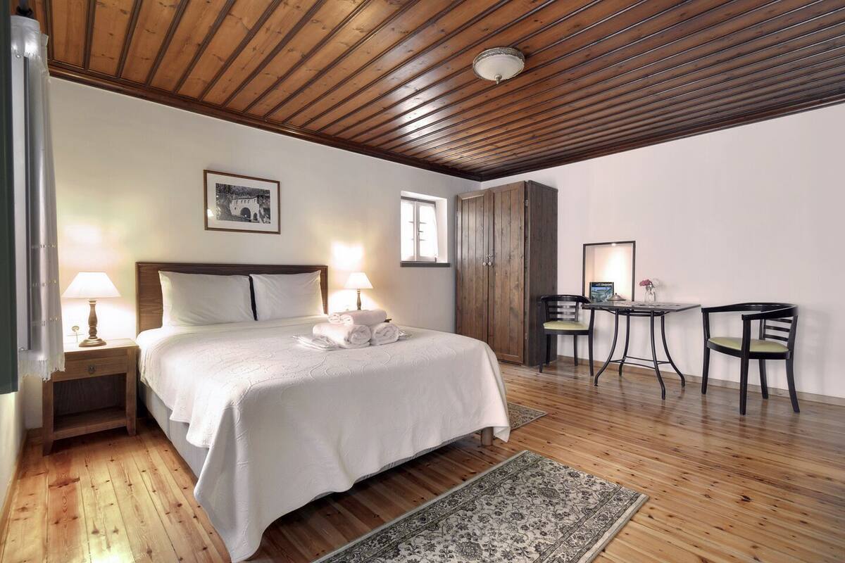 Ein Schlafzimmer mit Holzbalkendecke, einem Bett mit weißen Leinen, zwei Nachttischlampen, einem Nachttisch, einem Schreibtisch mit Stuhl und einem Fenster.