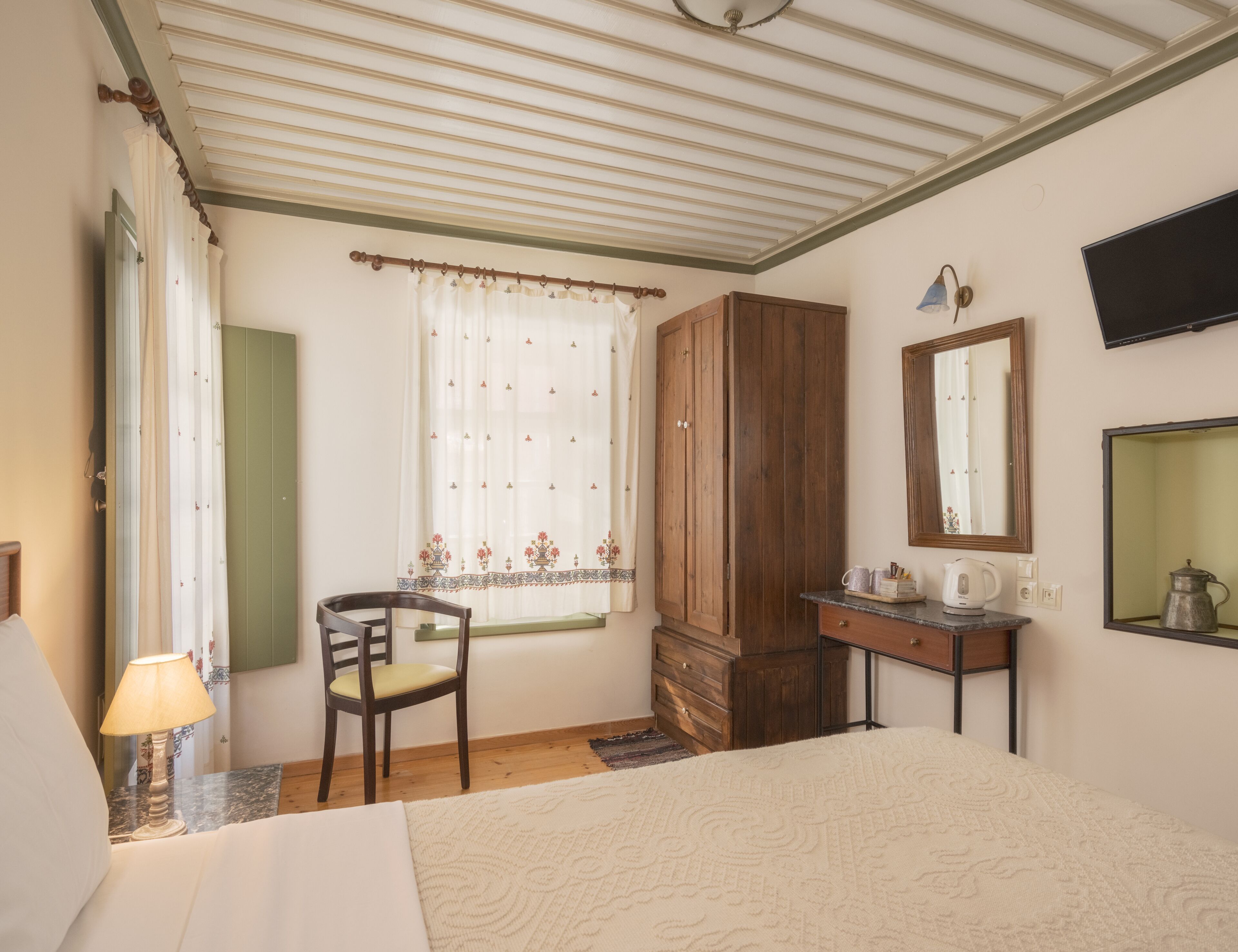 A bedroom with a bed, a wooden wardrobe, a desk with a mirror, a chair, and a window with curtains.