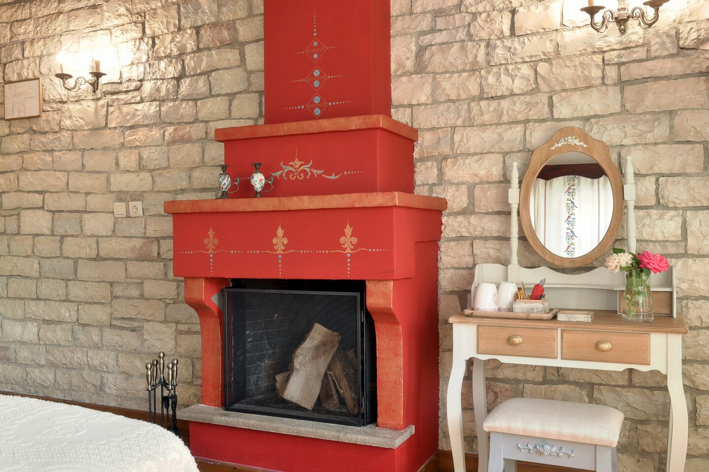 Una habitación con pared de piedra, chimenea roja con detalles decorativos, cómoda de madera con espejo y un banco.