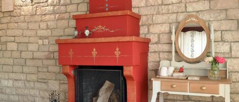 Una habitación con pared de piedra, chimenea roja con detalles decorativos, cómoda de madera con espejo y un banco.