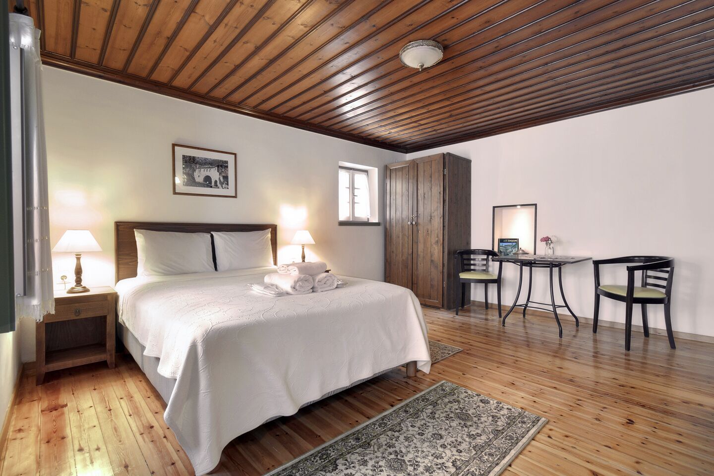 A bedroom with a wooden ceiling, a bed with white linens, two bedside lamps, a wooden wardrobe, a small table with a chair, and a patterned rug on the floor.