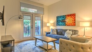 Una sala de estar con sofá, sillón, mesa de centro y lámparas. Hay una puerta corrediza de vidrio que da a un área exterior.