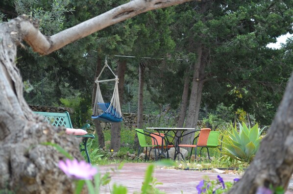 A hammock hanging between trees, a table with chairs, and a stone wall in a garden setting.