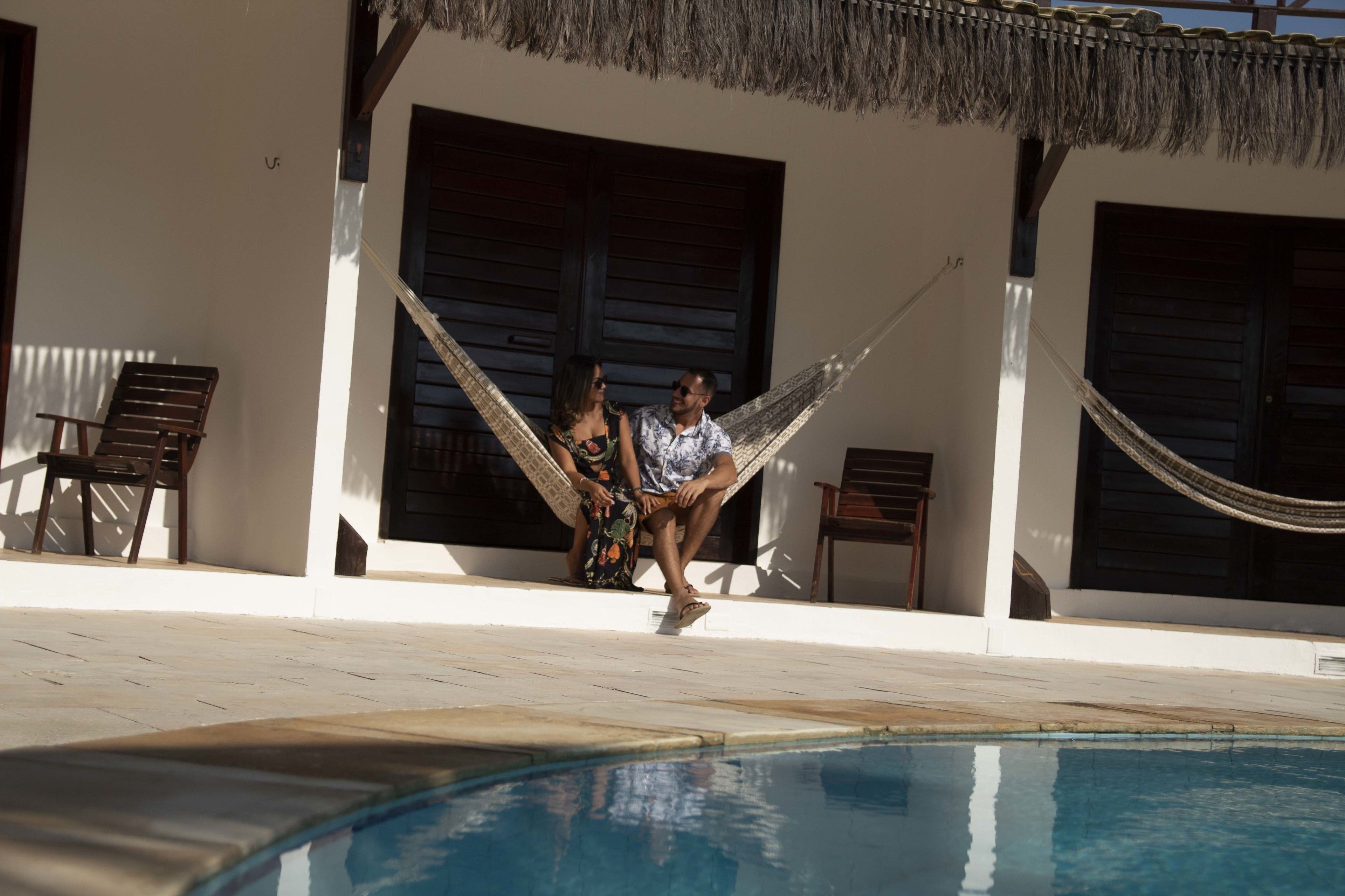 A couple sitting on a hammock by a poolside, with a clear view of the pool and a building with large windows in the background.