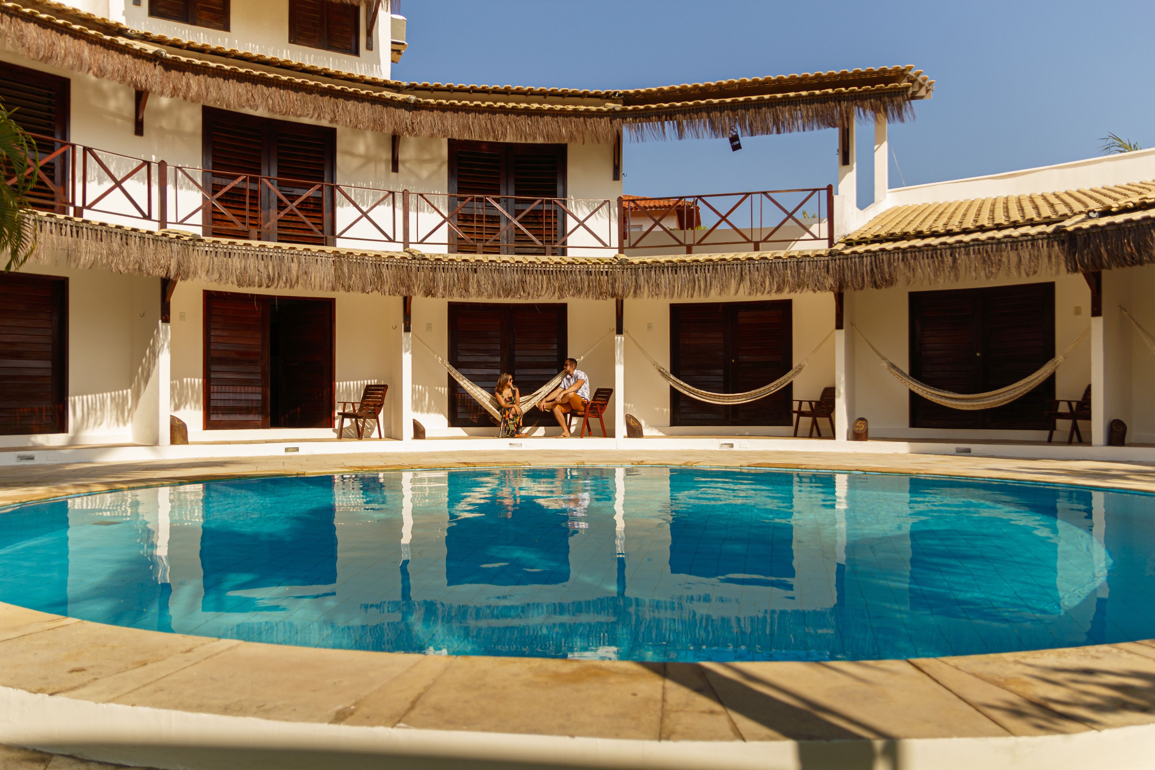 A pool area with a clear blue pool, a thatched-roof building, and a hammock.