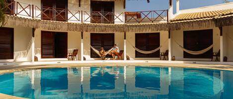 A pool area with a clear blue pool, a thatched-roof building, and a hammock.
