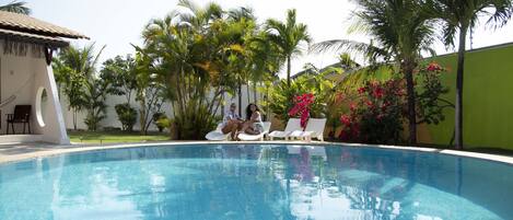 Una piscina con sillones de descanso, palmeras y un pequeño edificio con techo de paja.
