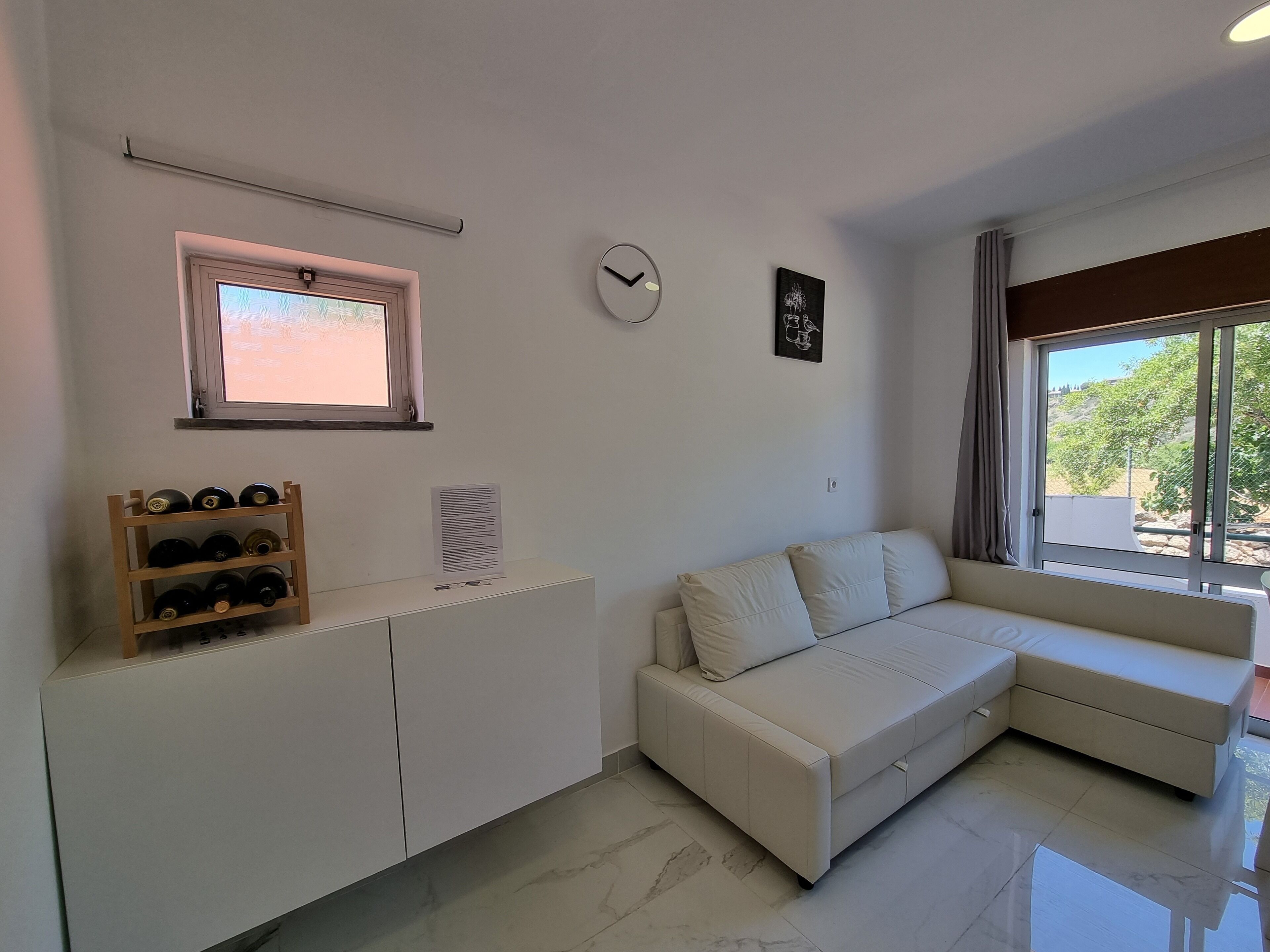 A modern living room with a white sofa, a wooden wine rack, a clock, and a window with a view of greenery.