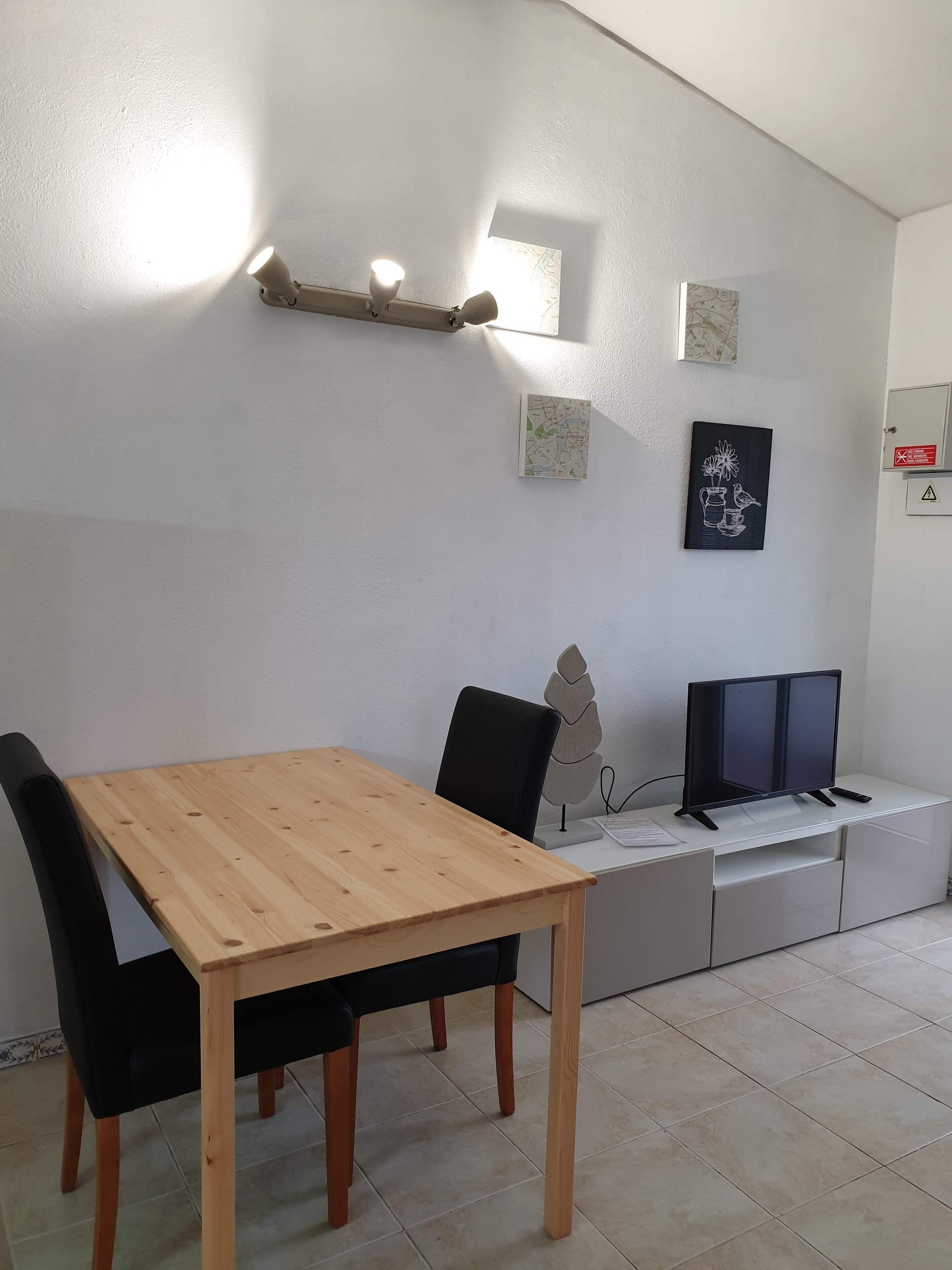 A modern living room with a wooden dining table, black chairs, a black sofa, a TV on a white cabinet, and wall-mounted lights.