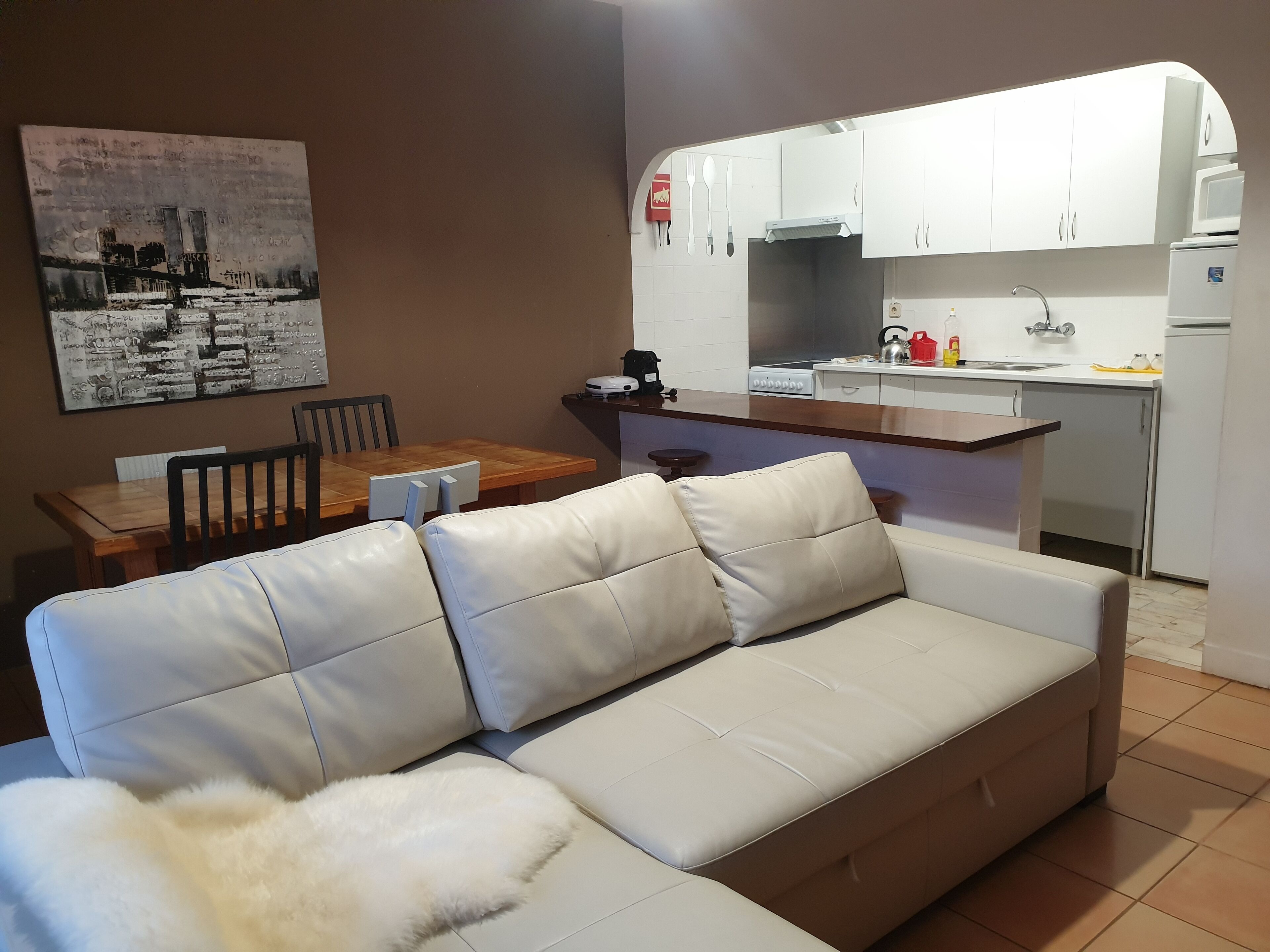 A modern living room with a white leather sofa, a dining table with chairs, and a kitchen area with white cabinets and appliances.