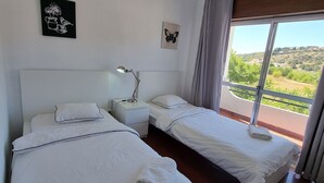 Hotellihuone, jossa on kaksi sänkyä, yöpöytä lampun kanssa, ikkuna, josta avautuu näkymä vehreään maisemaan, ja taideteoksia seinillä.