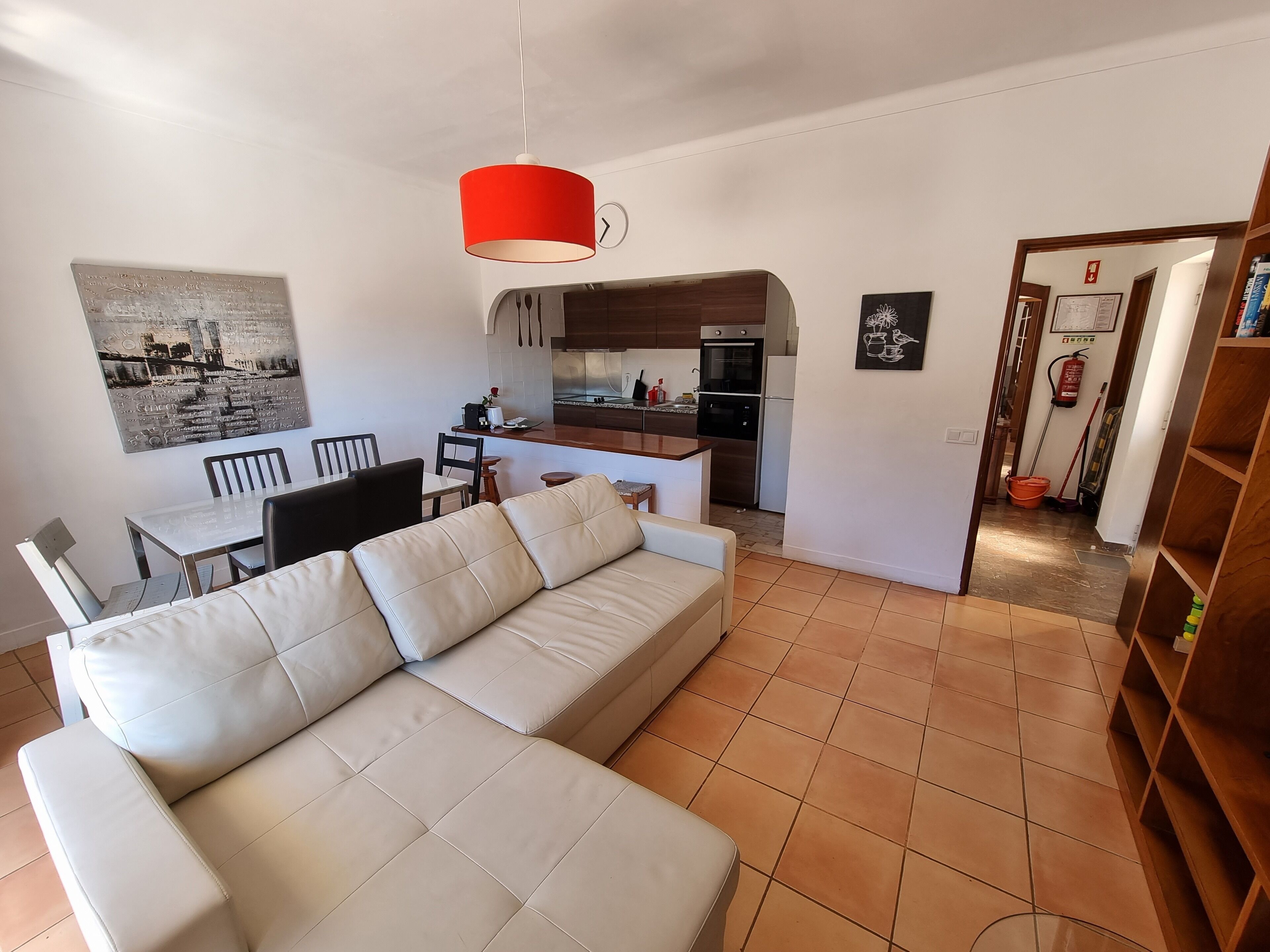 A modern living room with a white sofa, a dining area with chairs, a kitchen with a red hanging light, and a hallway with a fire extinguisher.