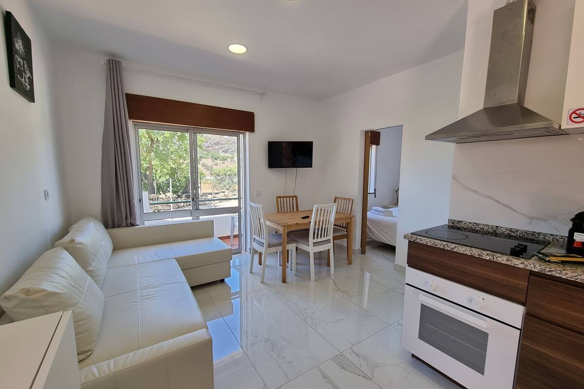 A modern living room with a sofa, dining table, and chairs. There is a kitchen area with a stove and oven. A TV is mounted on the wall, and a window provides a view of the outdoors.