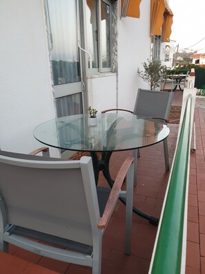 Una mesa con cubierta de vidrio al aire libre, con cuatro sillas, una planta en maceta y un banco verde.