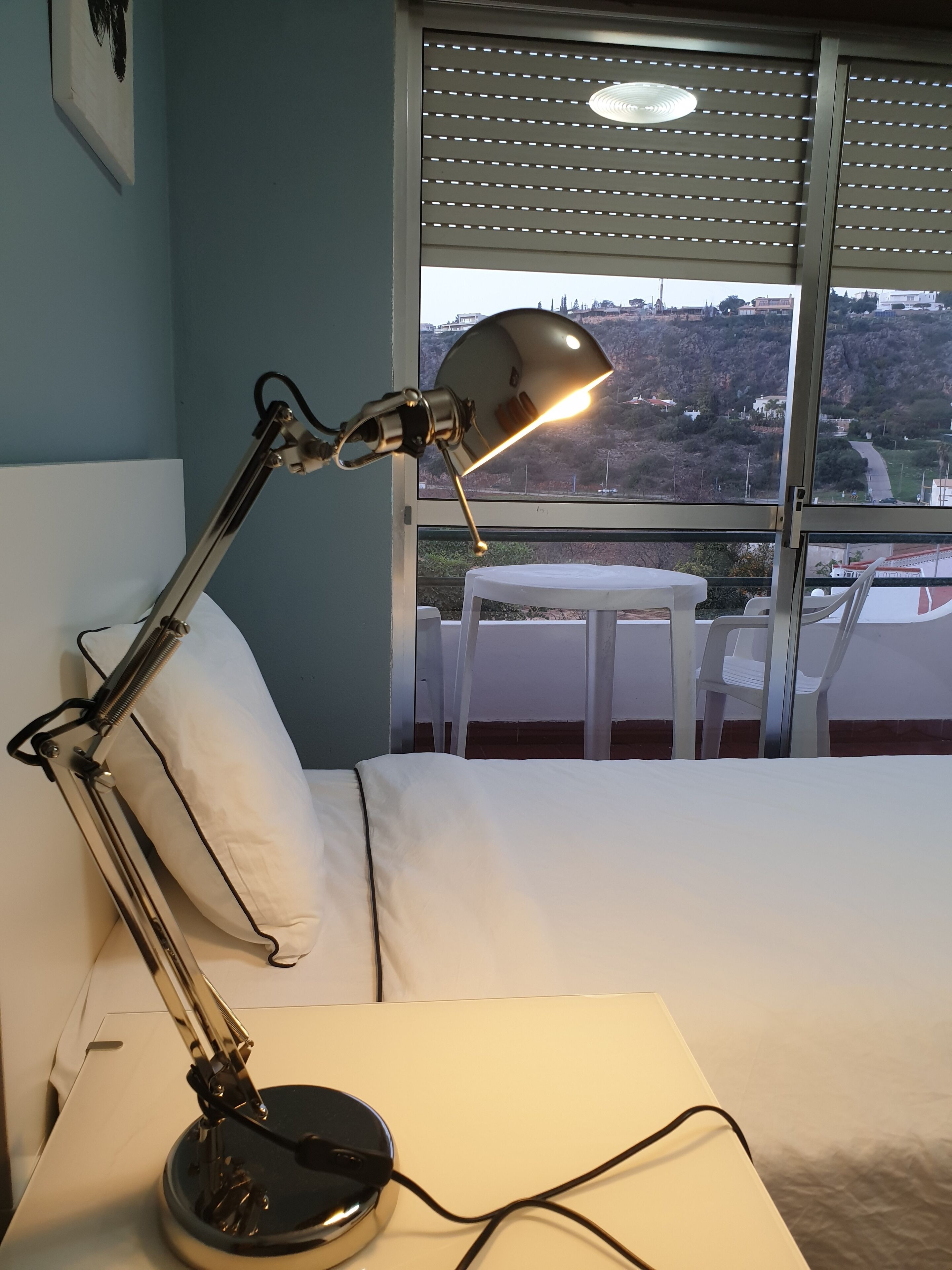 A hotel room with a bed, a desk lamp, a window with a view of a cityscape, and a small table with chairs.