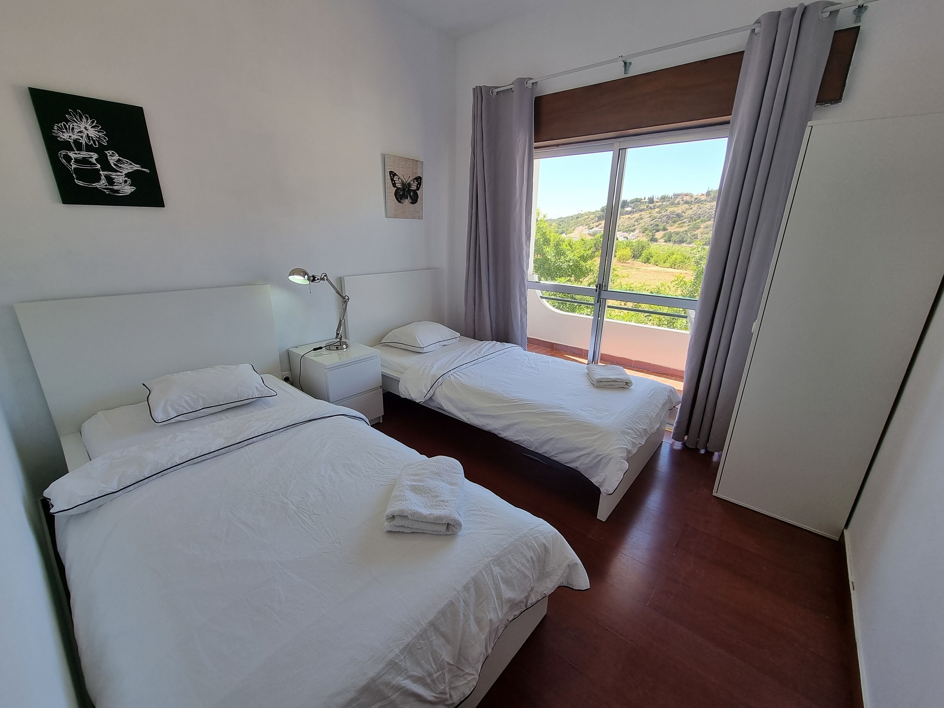 A hotel room with two beds, a nightstand with a lamp, a window with curtains, and artwork on the wall.