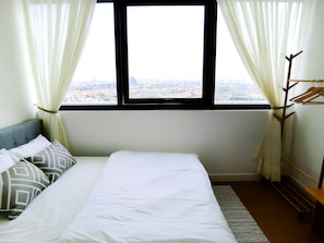 A hotel room with a bed, a window with curtains, a wooden hanger rack, and a view of a cityscape.