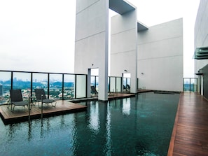 A rooftop infinity pool with wooden decking and lounge chairs, surrounded by large windows offering a city view.
