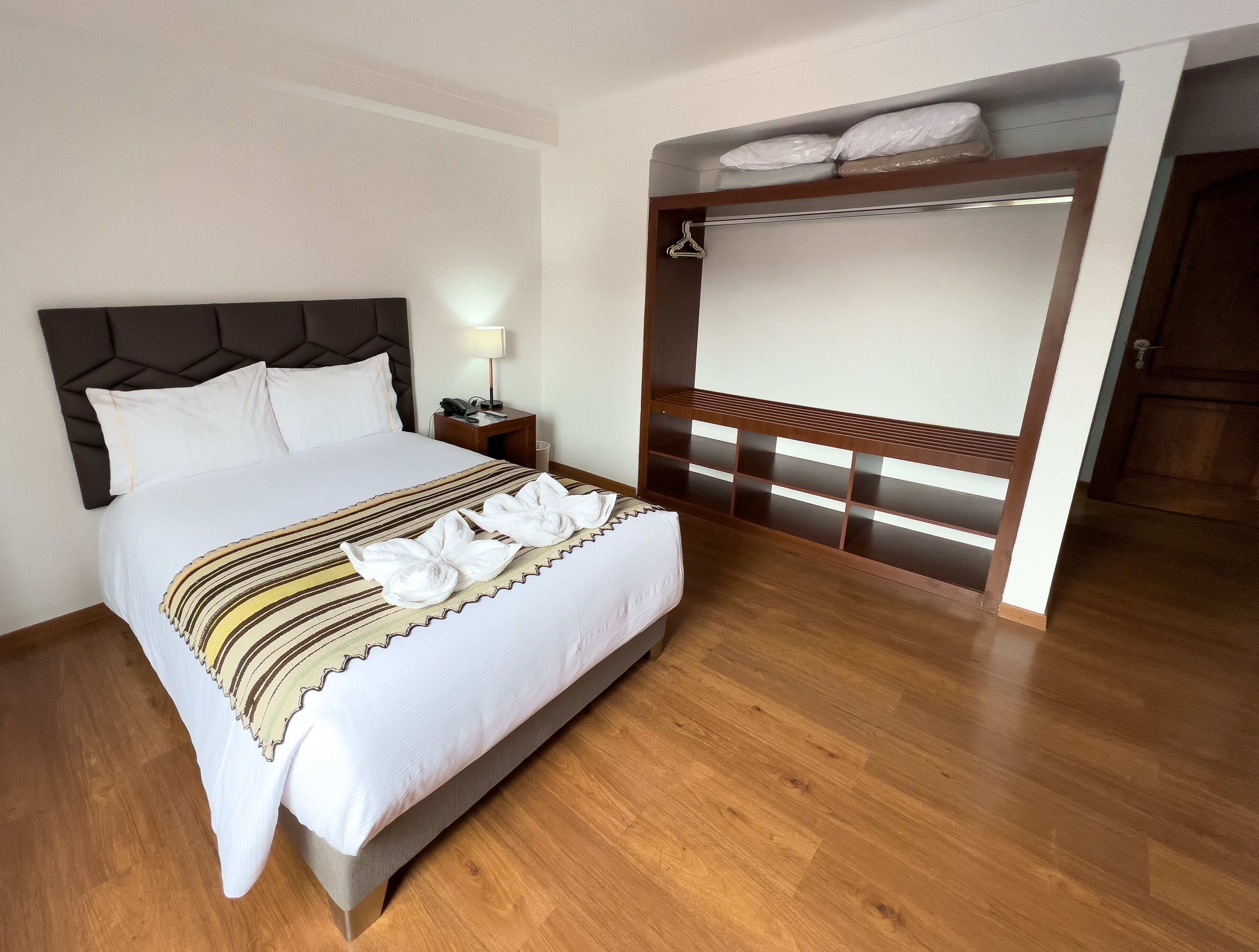 A hotel room with a large bed, a bedside table with a lamp, a wooden bench with towels, and a door leading to another room.