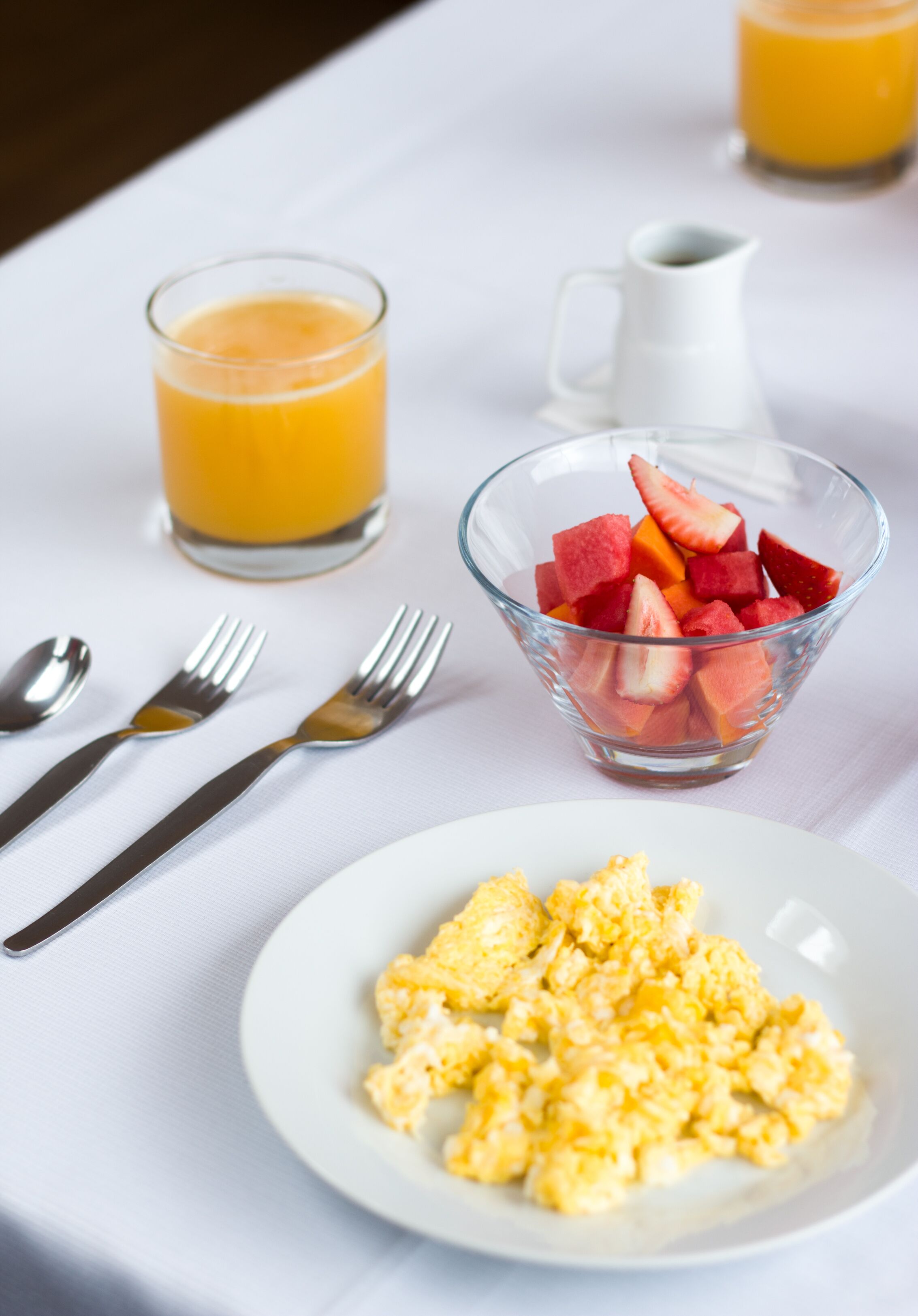 Desayuno con huevos revueltos, un tazón de fruta, un vaso de jugo de naranja y una jarra de agua sobre un mantel blanco.