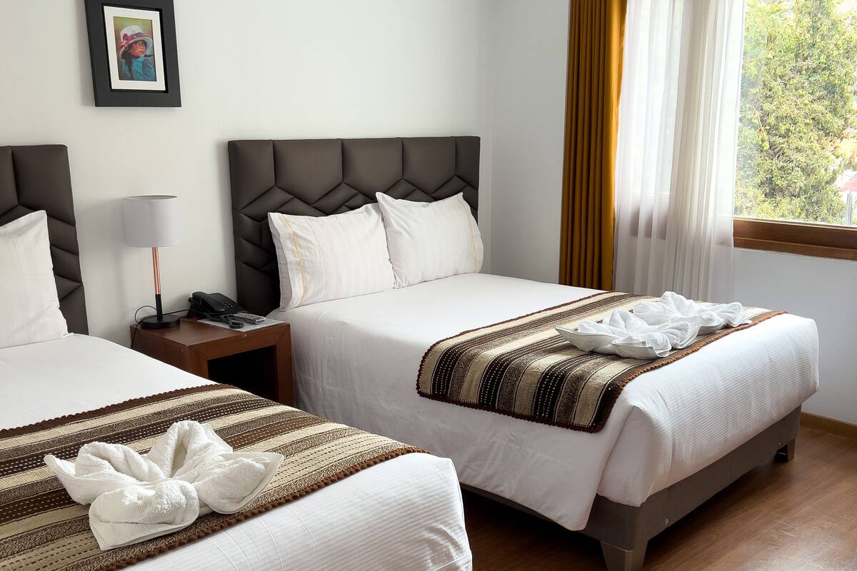 A hotel room with two beds, a bedside table with a lamp, a framed picture on the wall, and a window with curtains.