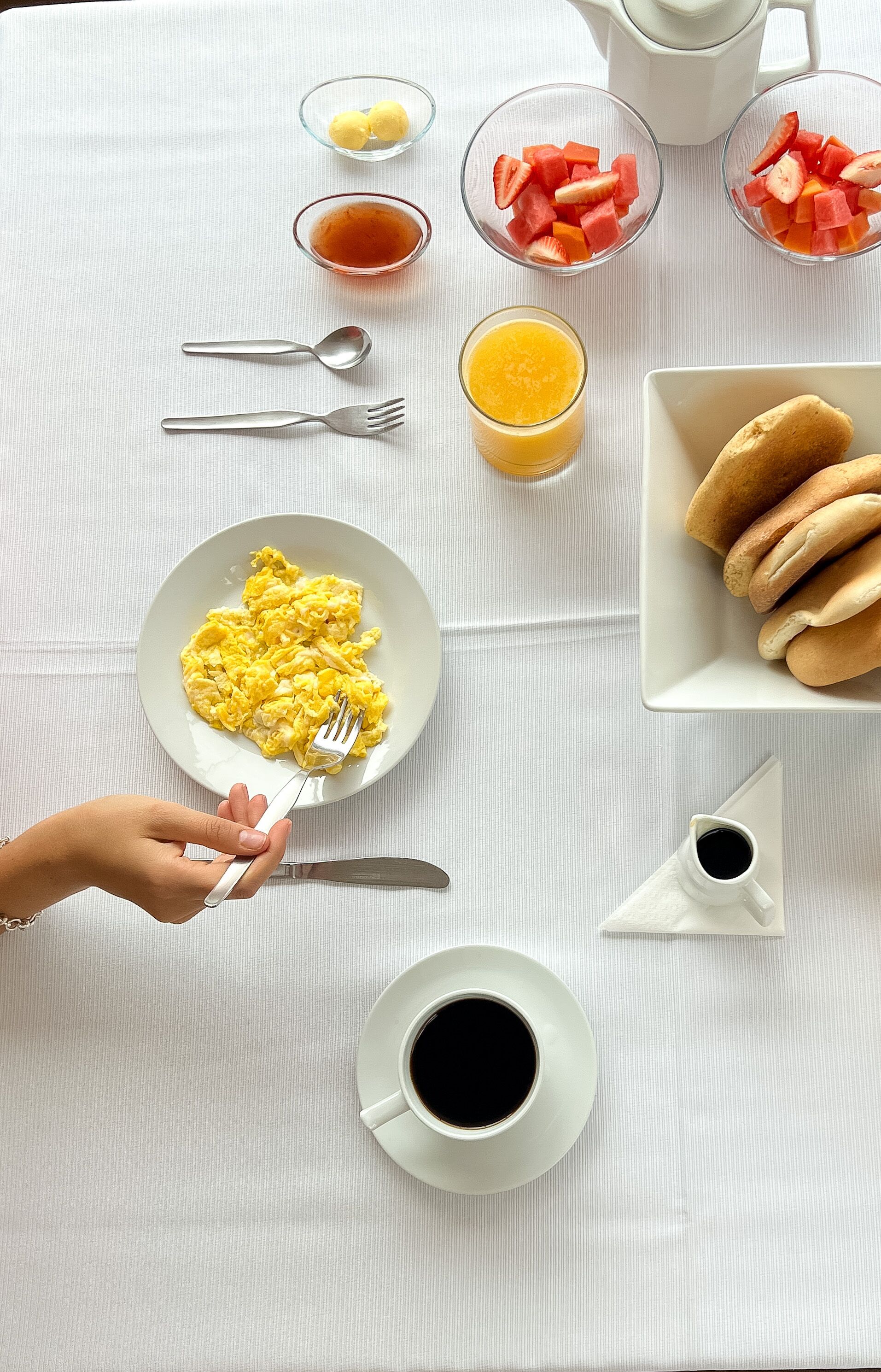 A breakfast setup with scrambled eggs, toast, fruit, and beverages on a white tablecloth.