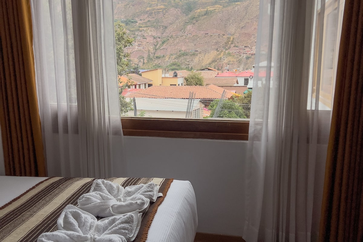 A hotel room with a large bed, a window with curtains, and a view of a hilly landscape.