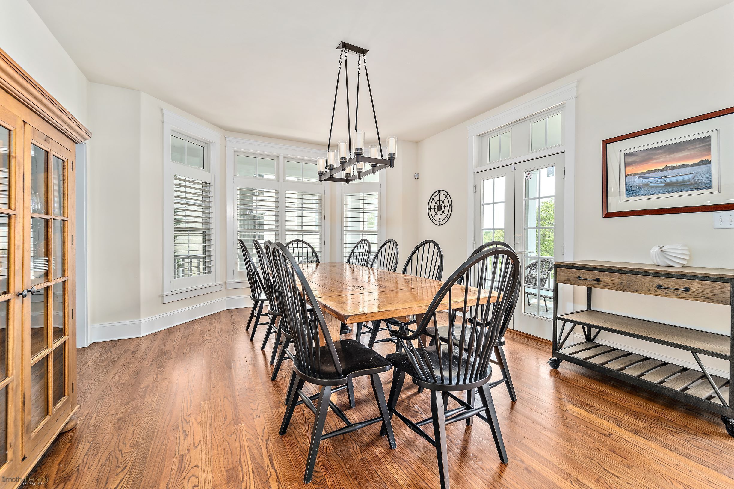 Une salle à manger avec une table et des chaises en bois, un lustre et une vue sur l’extérieur grâce à de grandes fenêtres.