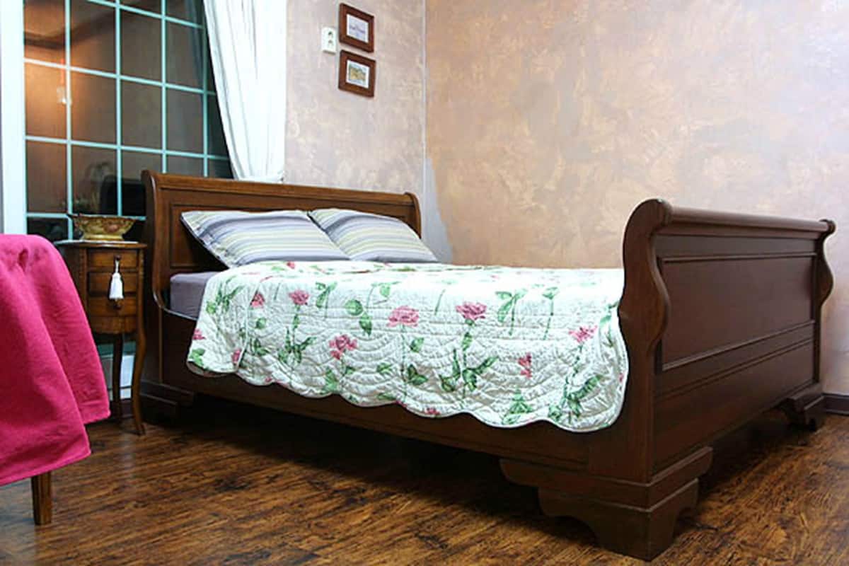 A wooden bed with a floral bedspread, a nightstand with a lamp, and a window with curtains.