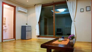 Una habitación con piso de madera, una mesa pequeña con un florero, un refrigerador y una puerta corrediza de vidrio que da a un balcón.