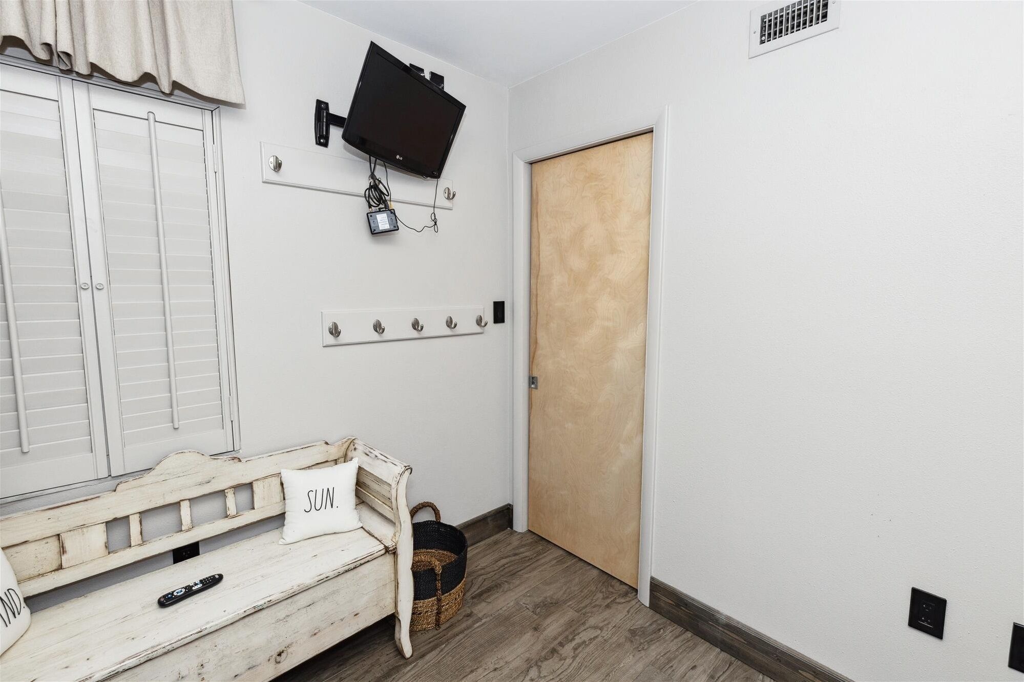 A room with a white wall, a wooden door, a white bench with a cushion, a mounted TV, and a wall-mounted light switch.