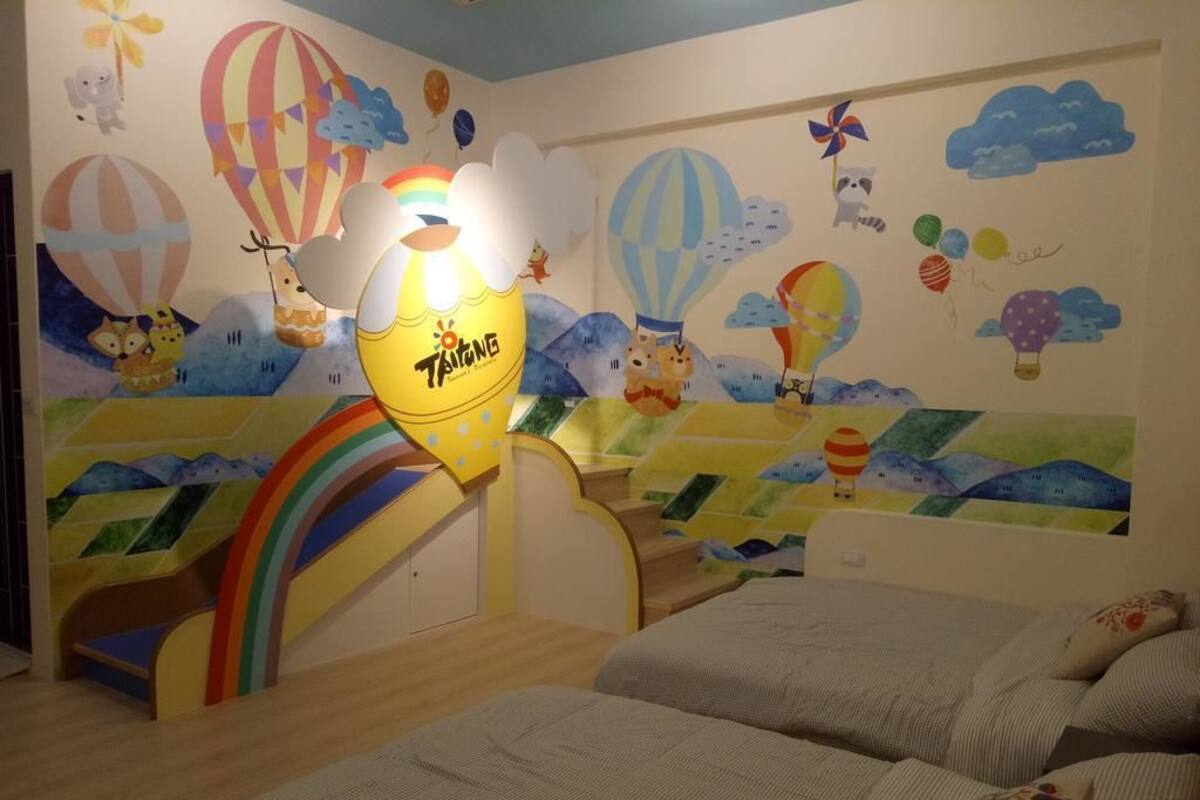 A child's bedroom with a bed, a small desk, and a colorful wall mural featuring hot air balloons, clouds, and a rainbow.