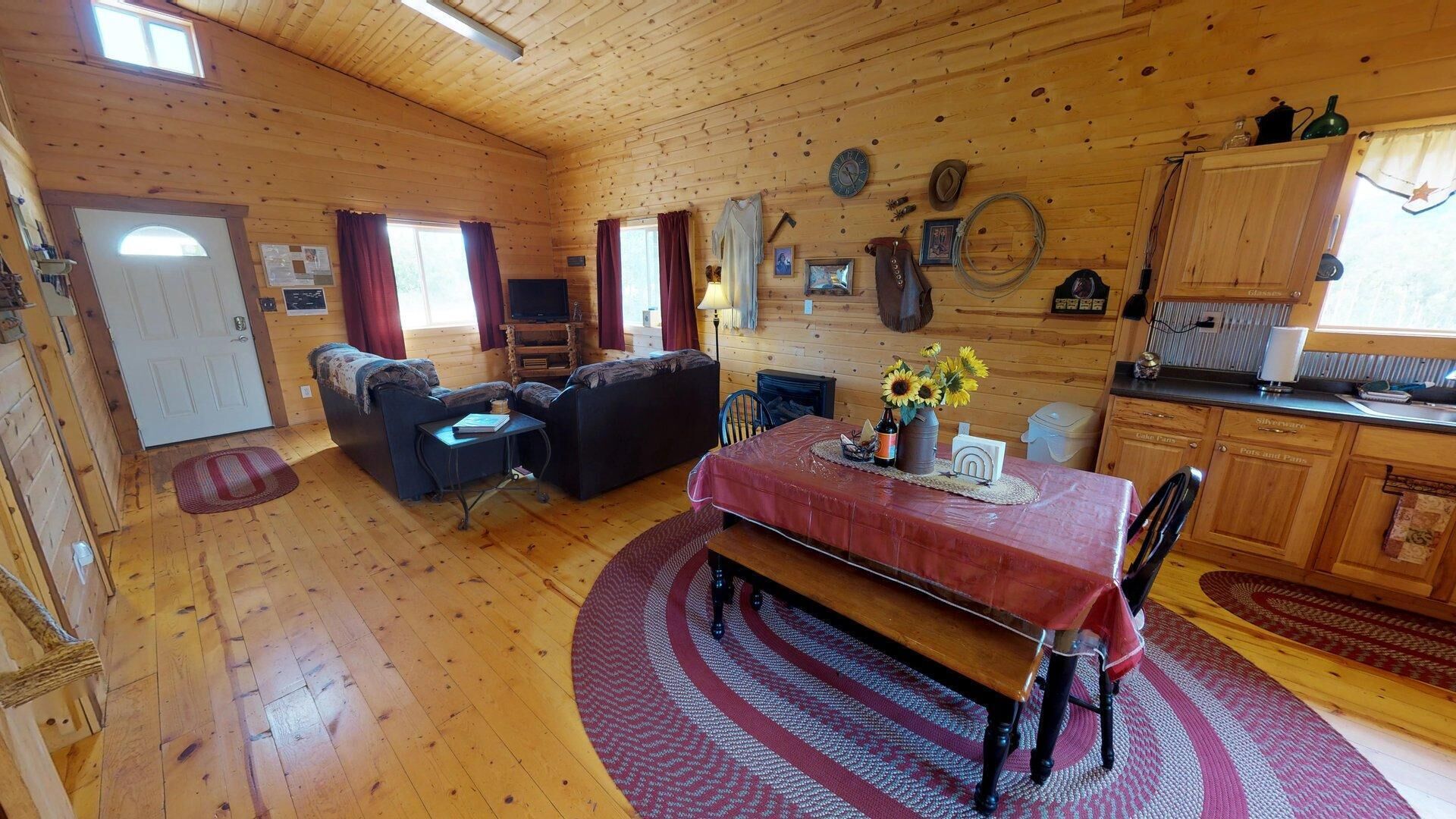 A cozy cabin interior with a dining area, a living room with a sofa and armchairs, wooden walls, and a kitchen area.