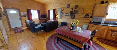 A cozy cabin interior with a dining area, a living room with a sofa and armchairs, wooden walls, and a kitchen area.