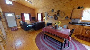 A cozy cabin interior with a dining area, a living room with a sofa and armchairs, wooden walls, and a kitchen area.