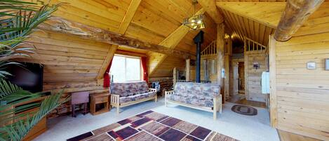 A cozy log cabin interior with a wooden ceiling, a sofa set, a television, and a large window.