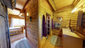 A wooden cabin interior with a bed, a bathroom with a sink and mirror, and a window with curtains. - Canyon View Family Lodge (Monticello)