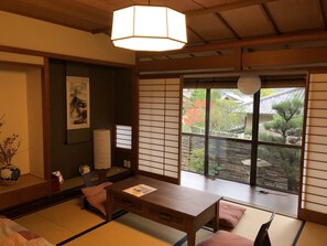 A traditional Japanese room with tatami flooring, a low wooden table, a hanging light fixture, and a view of a garden through a sliding door. - Kinkaku Ryokan (Kyoto)