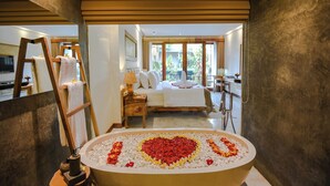 A bedroom with a large bed, a bathroom with a heart-shaped flower arrangement in the tub, and a view of the outdoors. - Purana Suite Ubud (Ubud)