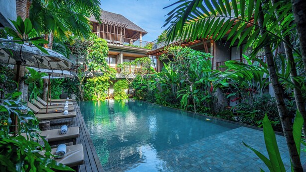 A pool area with lounge chairs and a wooden deck, surrounded by lush greenery and a building with a balcony. - Purana Suite Ubud (Ubud)