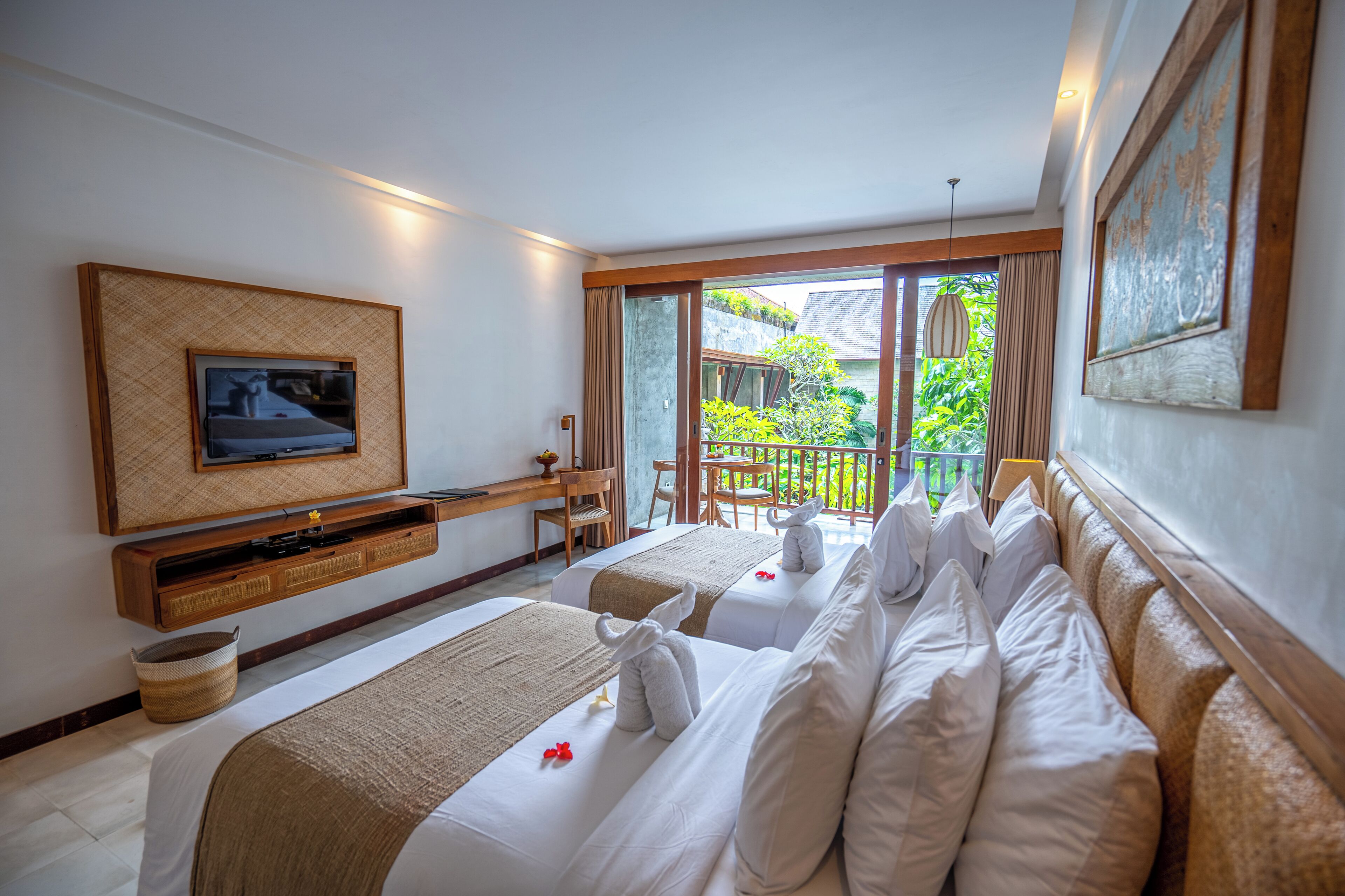 A hotel room with two beds, a TV, a desk, and a balcony with a view of greenery.