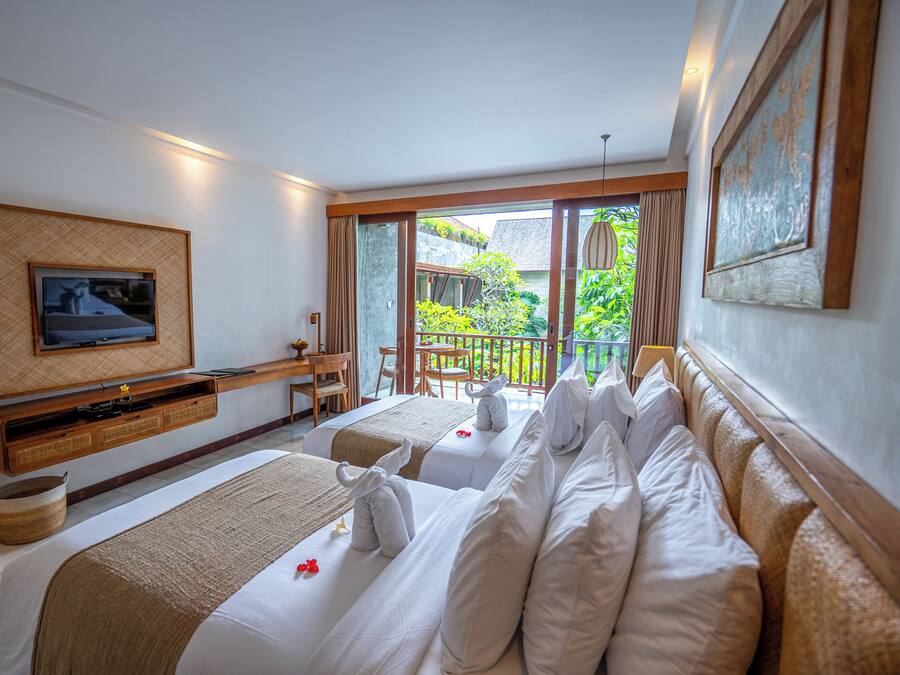 A hotel room with two beds, a TV, a desk, and a balcony with a view of greenery.