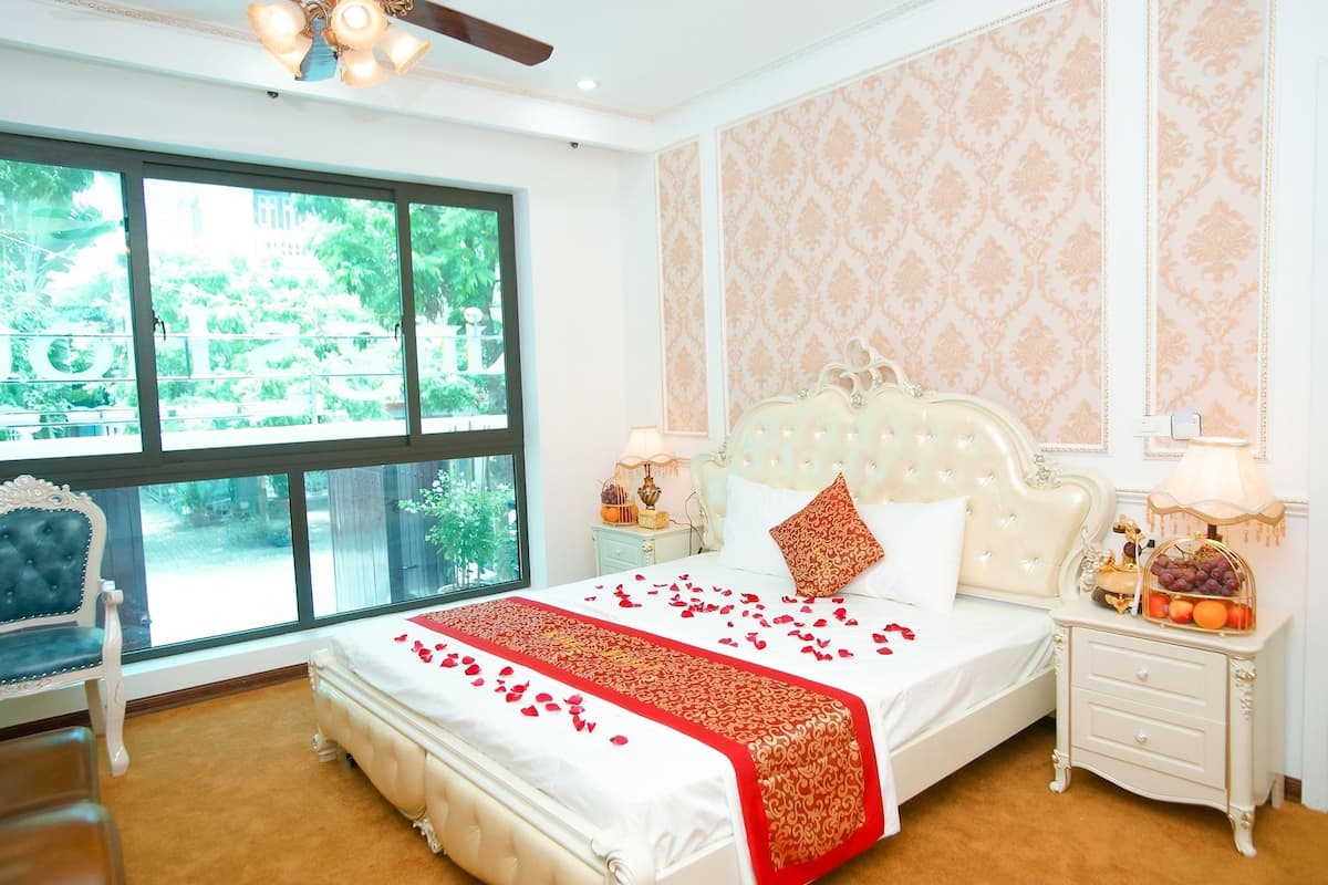 A bedroom with a large window, a bed with rose petals, a nightstand with fruit, and a chair.