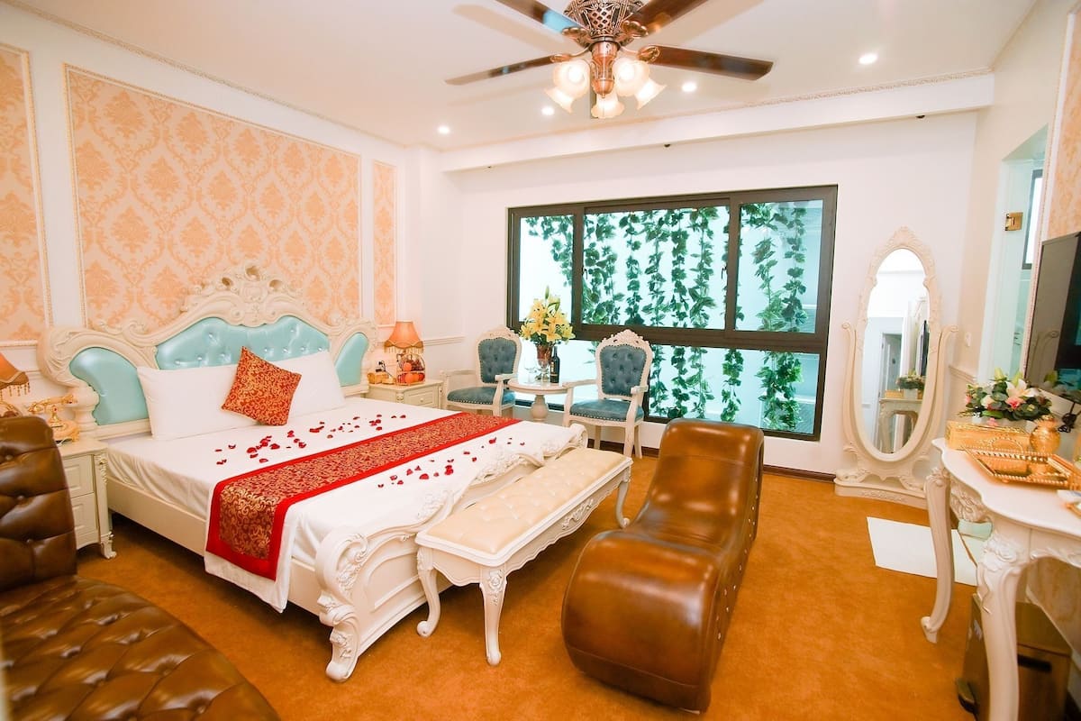 A bedroom with a bed, a sitting area with a chair, a vanity with a mirror, and a large window with a view of greenery.