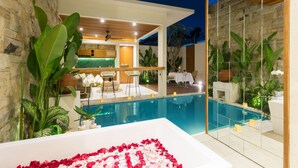 Uma área moderna de piscina interna, com parede de pedra, grandes plantas verdes e um espaço de refeições com mobiliário branco.