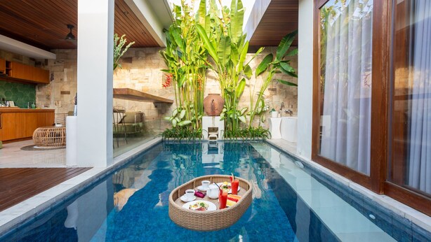 Uma piscina com uma bandeja flutuante de comida e bebidas, rodeada por plantas tropicais exuberantes e uma parede de pedra.