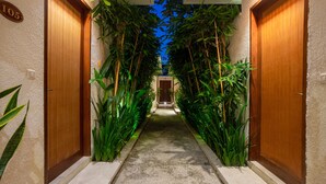 Corredor com portas de madeira, número 105, ladeado por plantas verdes altas, levando a uma porta no final.